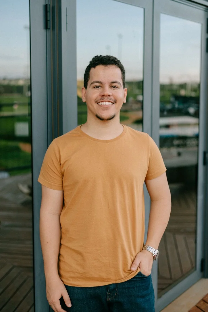 Nathan Nóbrega sorrindo, de camiseta laranja, em frente a uma porta de vidro